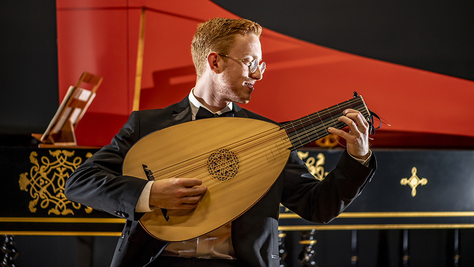 A student playing the guitar, sat in front of a covered harpsichord