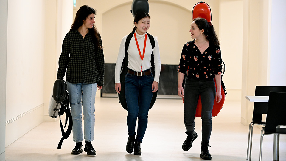 Three students carrying instrument cases walking through the RCM cafe