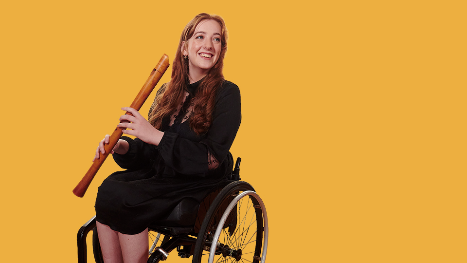 A women wearing a black dress, smiling, holding a historical clarinet, sitting in a wheelchair. A women wearing a black dress, smiling, holding a historical clarinet, sitting in a wheelchair.