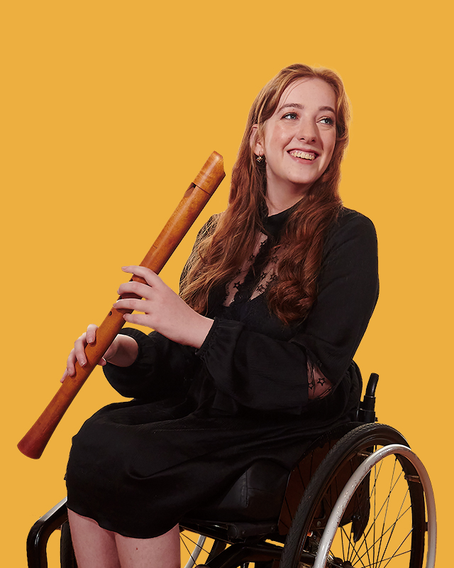A women wearing a black dress, smiling,  holding a historical clarinet, sitting in a wheelchair.
