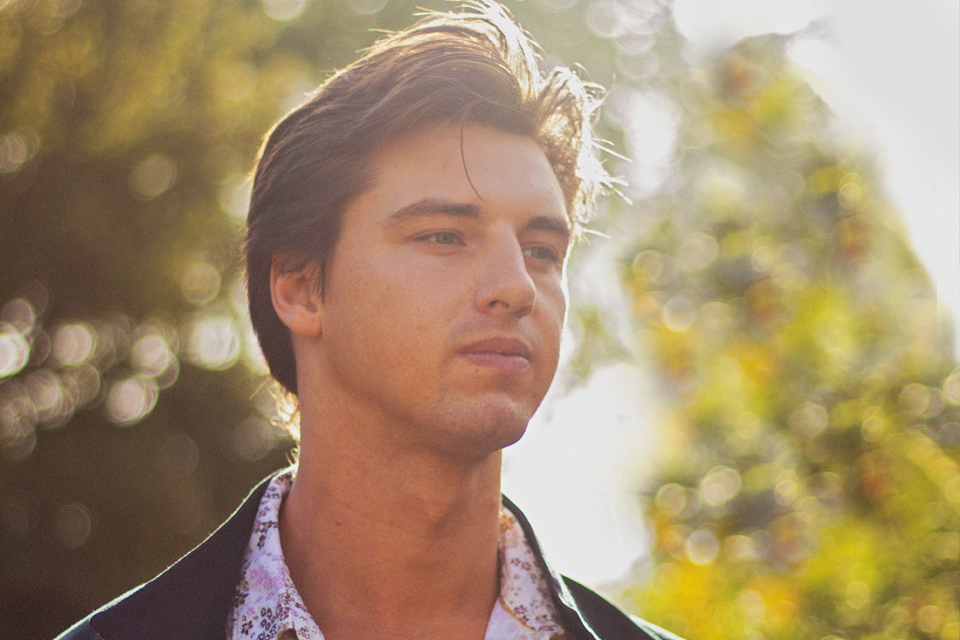 A man with brown hair, with a patterned shirt, wearing a black blazar, with a green trees in the background.