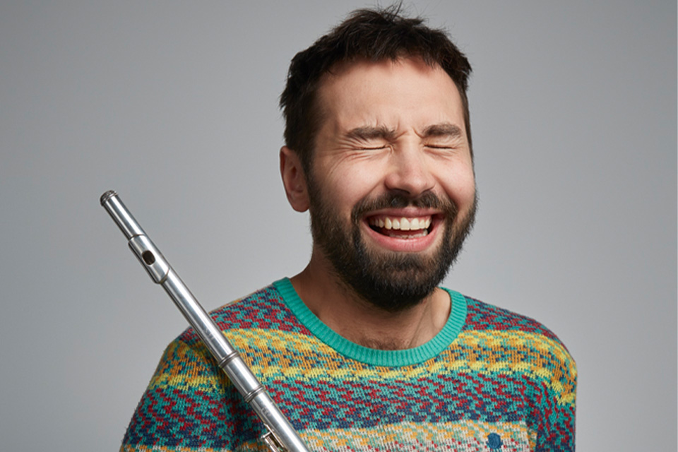 Adam Walker wears a green jumper and holds his flute, laughing