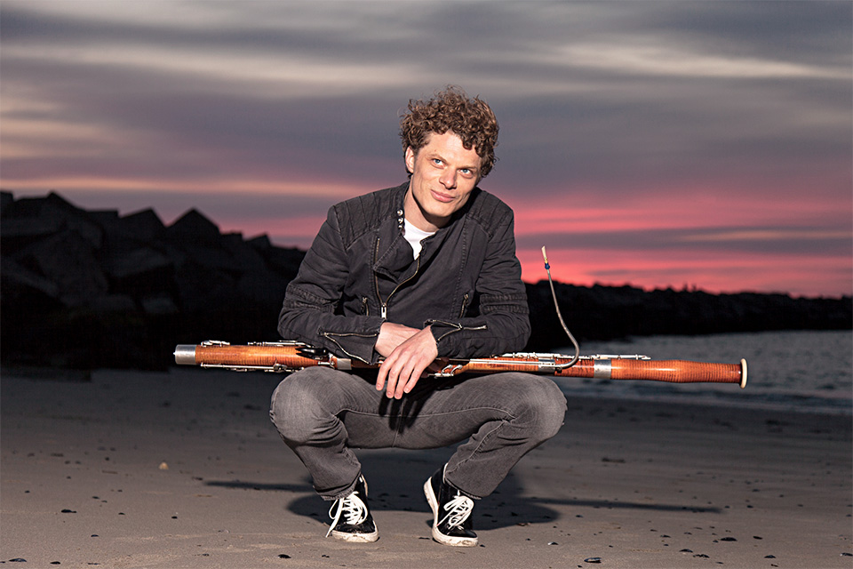 Bram van Sambeek crouches holding his bassoon, a pink sunset behind him