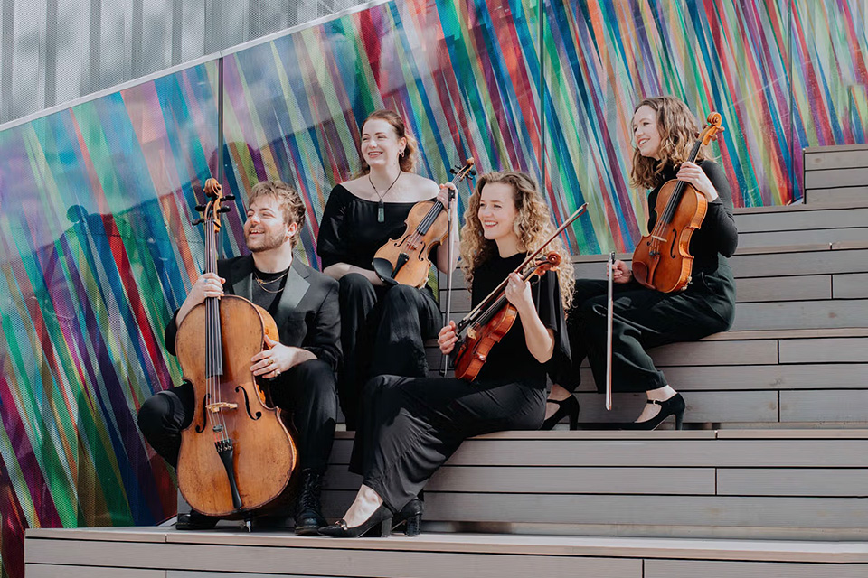The Calathea Quartet sit on steps outside smiling and holding their instruments