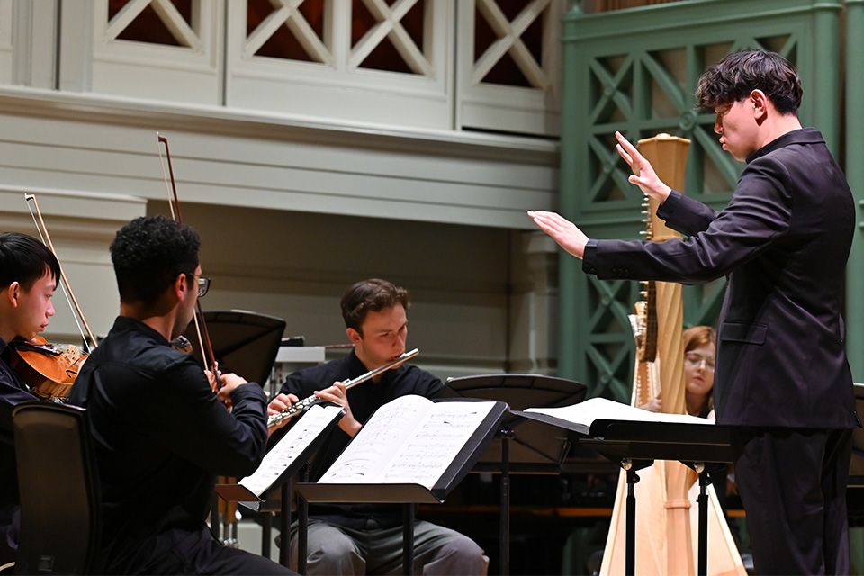 A man in a black shirt conducts a chamber group of violin, viola, flute and harp in the Performance Hall