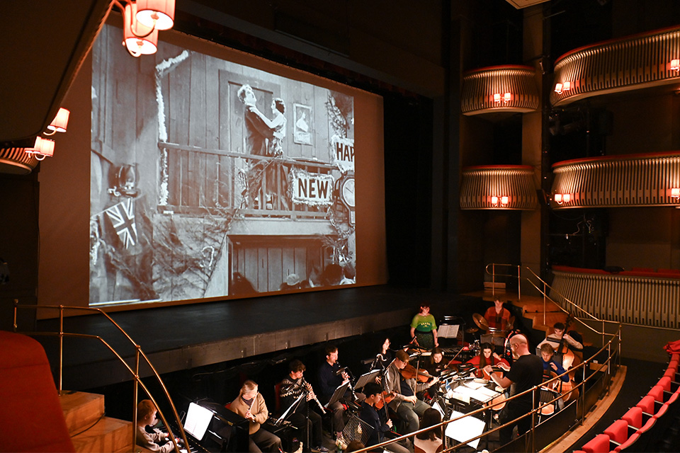 A conductor leads an orchestra in the pit of the Britten Theatre while a film is projected on a large screen above them