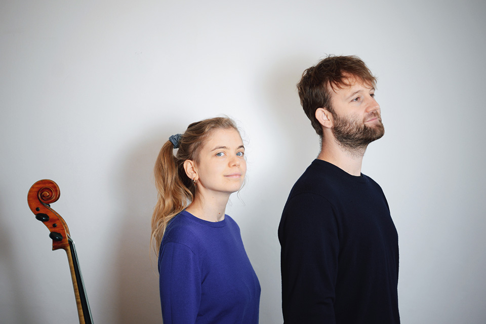 Héloïse Werner and Colin Alexander stand in front of a light grey wall wearing blue and black respectively