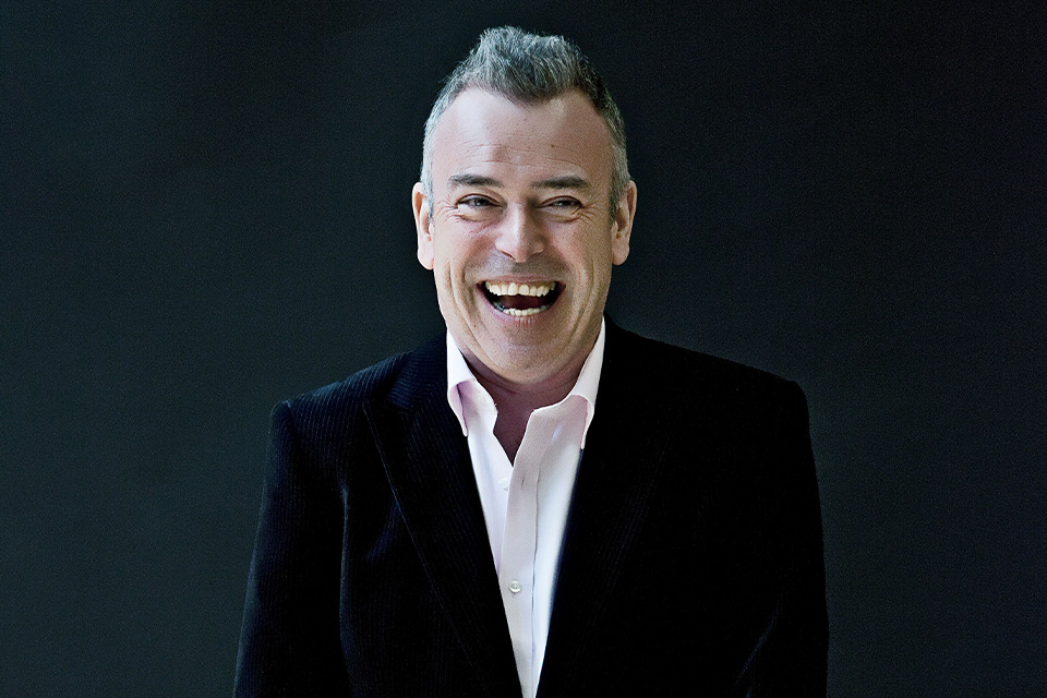 Malcolm Martineau wears a white shirt and black blazer and laughs broadly