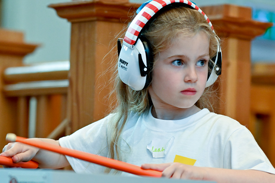 A young girl wears headphones and holds drum mallets