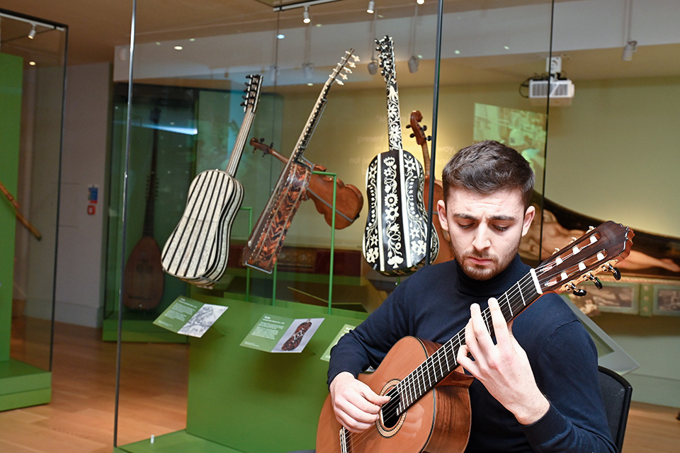 A man plays the guitar in the RCM Museum, a display case of stringed instruments behind him