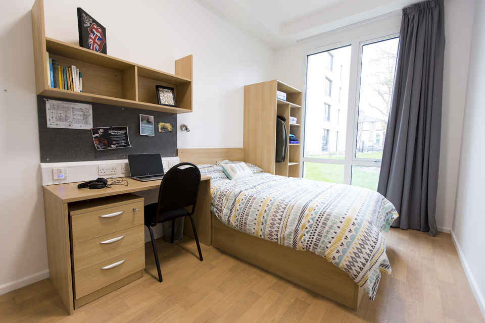 A room at the PCV, a single bed, with a desk next tot he bed, with shelves above and a large window showing the outside.