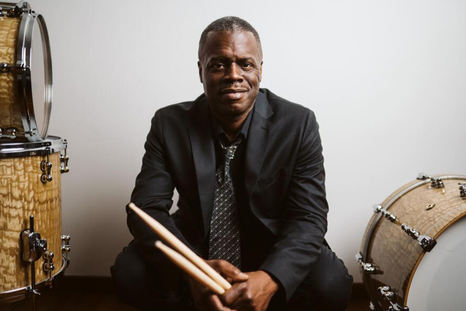 A man wearing a black suit, shirt and dark tie, holding a pair of drum sticks, smiling at the camera.