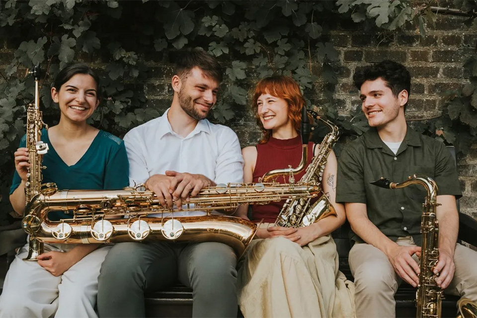 Versa Winds sit on a bench in front of a brick wall, holding their instruments and smiling