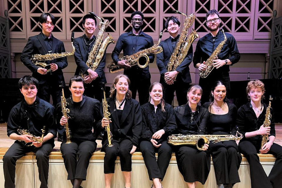 The RCM Saxophone Ensemble sit on the edge of the stage, holding their instruments and smiling