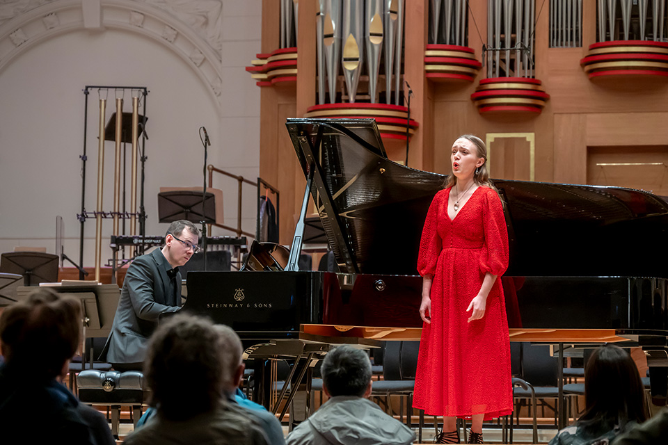 A singer wearing a red dress performs on stage, a pianist accompanying behind her