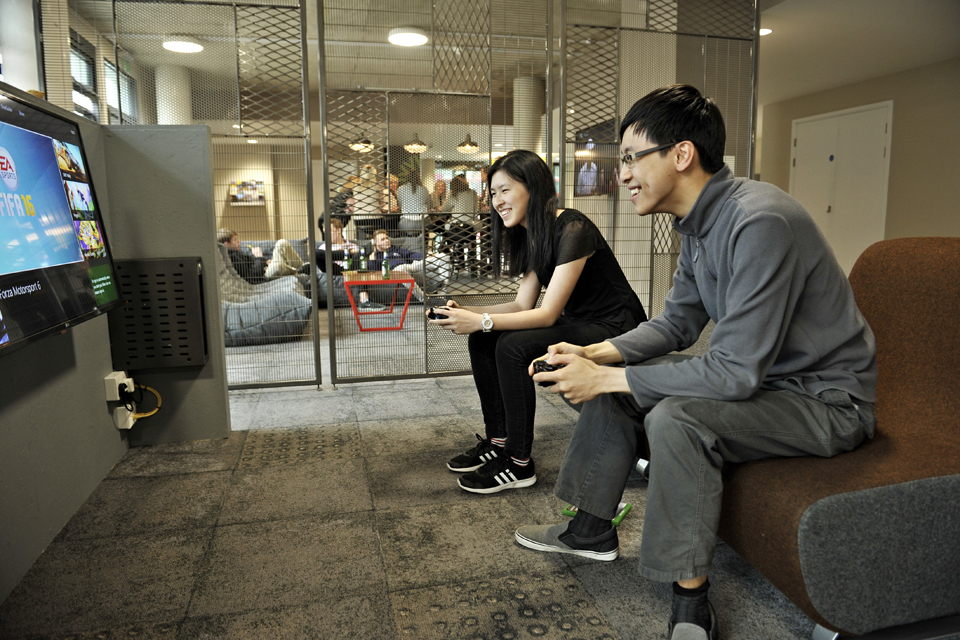 Two Asian students playing a video game, facing a screen with a game on it, sitting on a sofa together.
