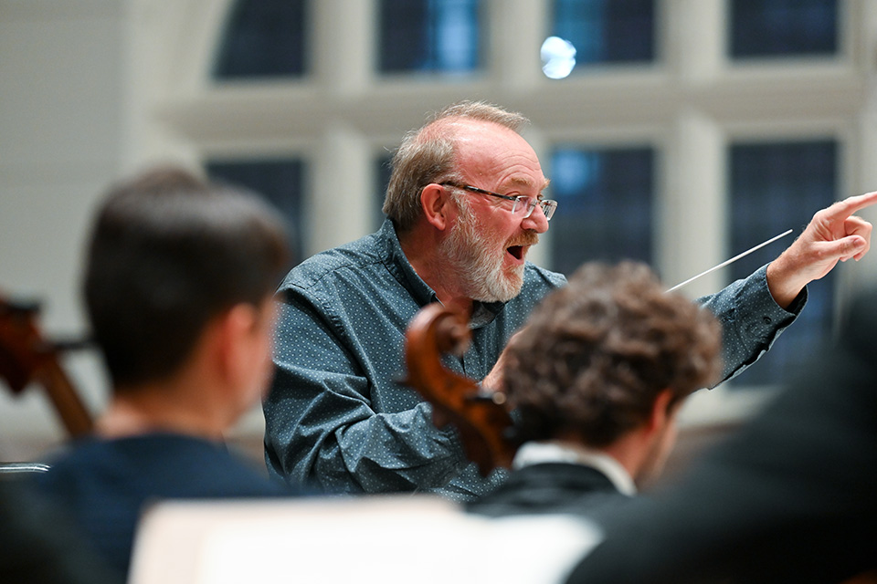 Martyn Brabbins conducts an orchestra, holding his baton and pointing animatedly
