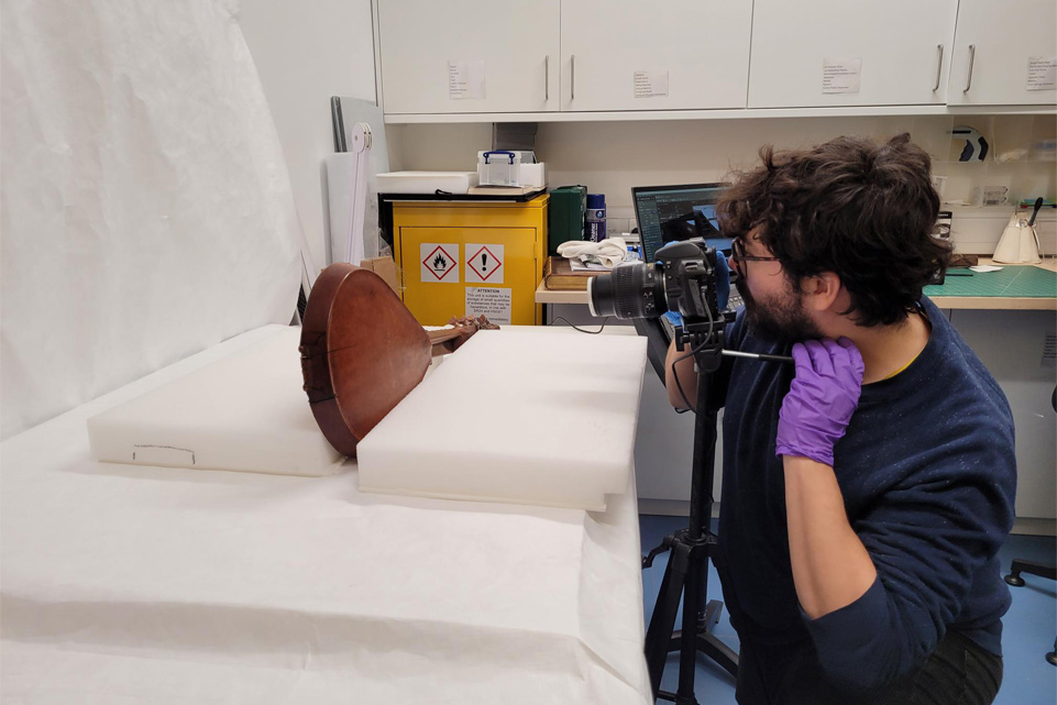 A male researcher takes photographs of a historic musical instrument, in front of a white background, in the museum research centre.