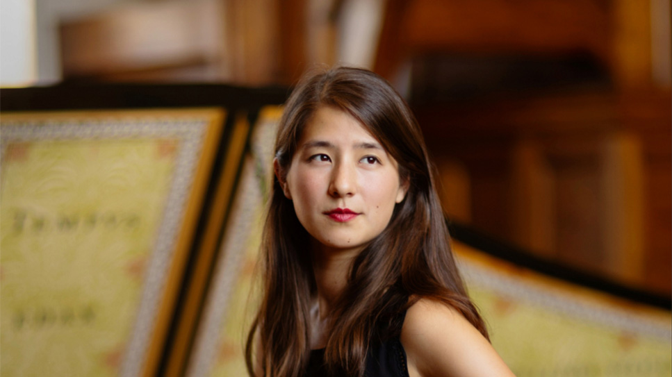 A women with long hair, sitting next to a harpsichord, looking away from the camera with a small smile. A women with long hair, sitting next to a harpsichord, looking away from the camera with a small smile.