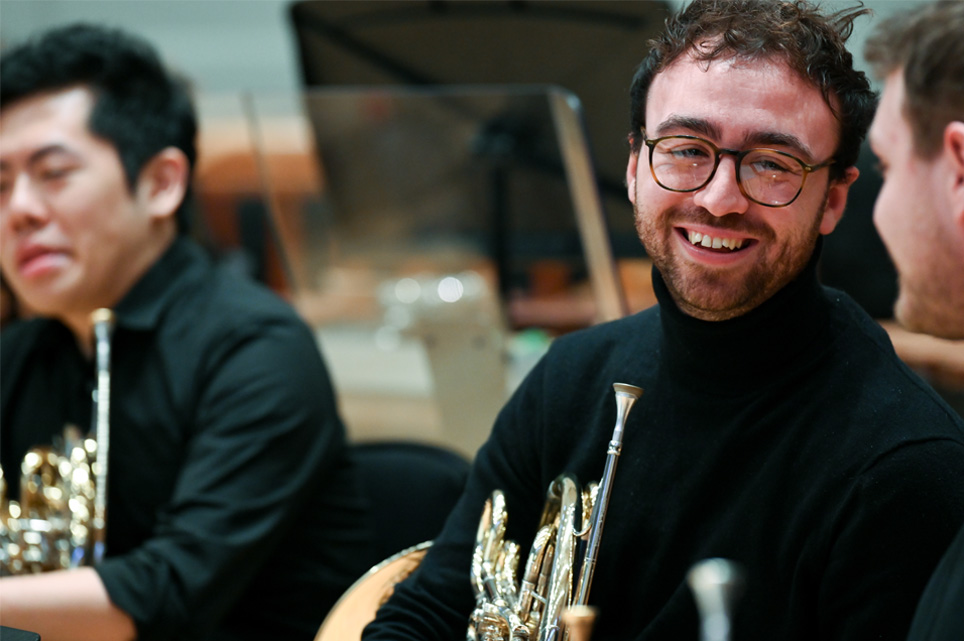A male student, wearing a black shirt, smiling and chatting to other students, all sitting down and holding their French horn, in a rehearsal setting. A male student, wearing a black shirt, smiling and chatting to other students, all sitting down and holding their French horn, in a rehearsal setting.