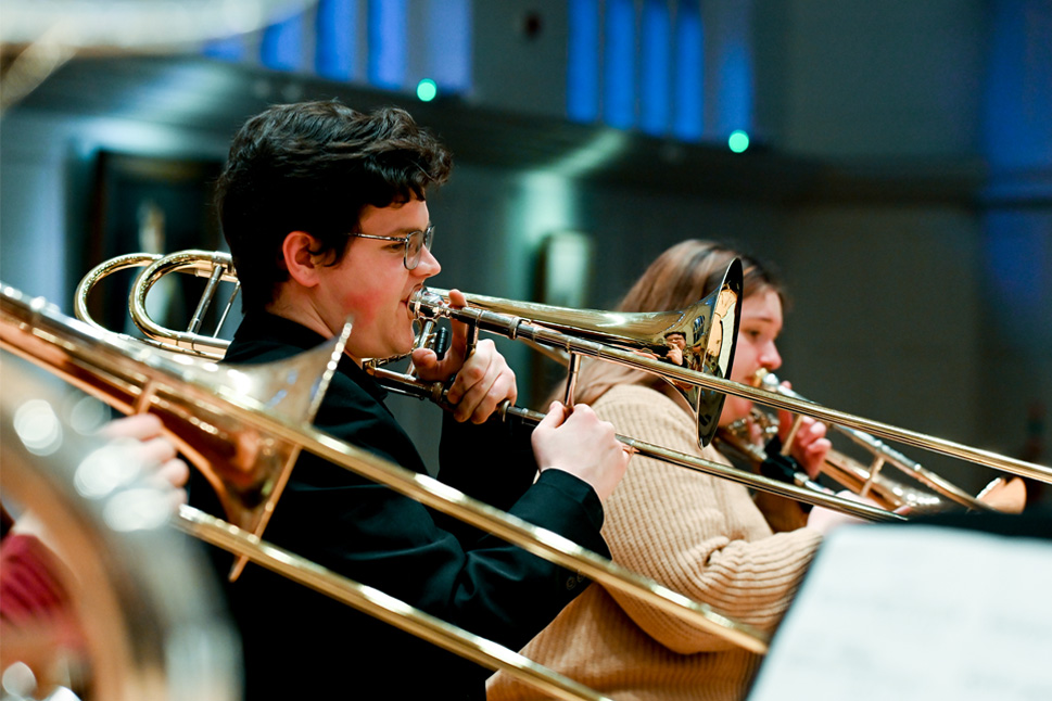 A group of trombonists, focusing on one male trombone player, wearing a black top, performing together in a rehearsal in a performance space
