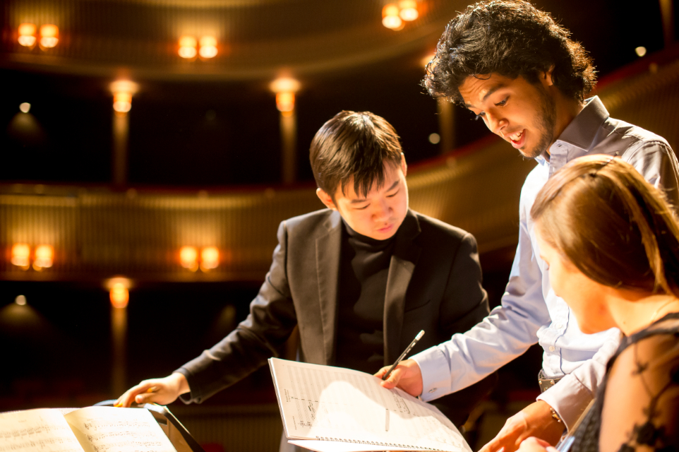 A student, holding a composition music sheets, talking to two other students, who are looking at the sheets, on the stage of a well lit theatre.