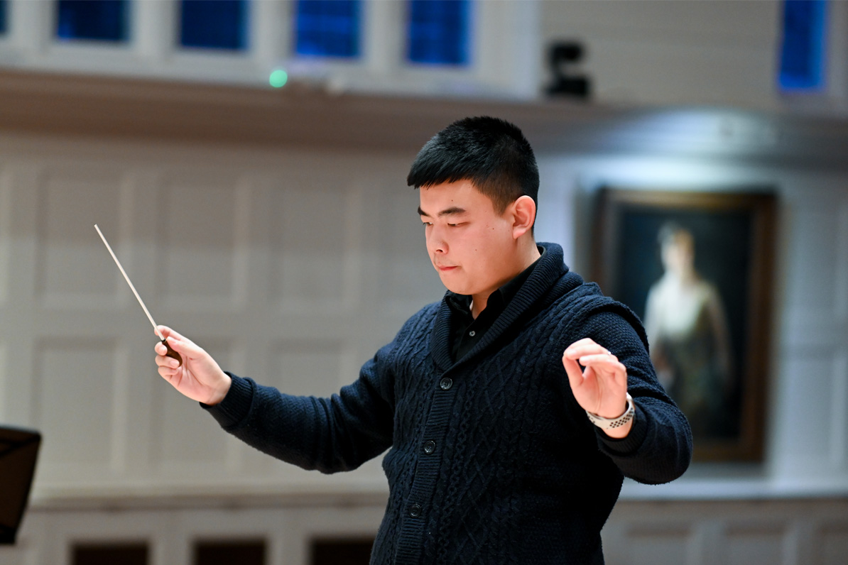 A male student, wearing a smart black shirt, conducting a brass ensemble in a performance room. A male student, wearing a smart black shirt, conducting a brass ensemble in a performance room.