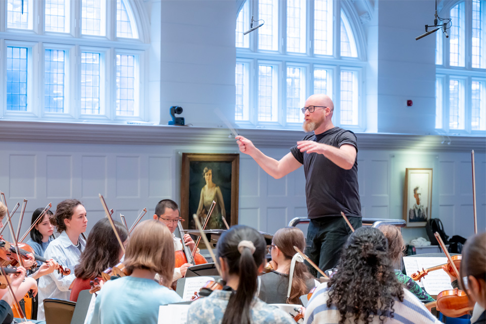 A man, wearing a dark shirt and glasses, conducting an orchestra rehearsal with students performing in a well lit concert hall.