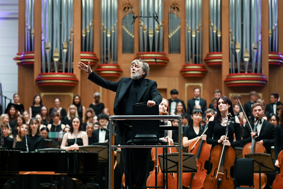 A man, wearing a black blazar and black shirt, standing in the podium, waving to the audience, with a group of students standing and holding their instruments, behind him.