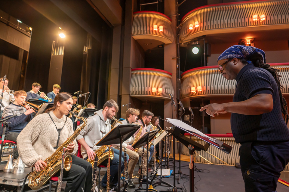 A male conductor, wearing a hair wrap, wearing a dark blue shirt, conducting a group of students performing in a jazz orchestra on a stage.