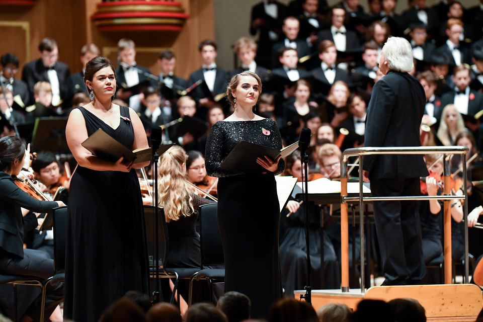 Two female singers, in smart black dresses, standing on stage, with an orchestra performing in the background.