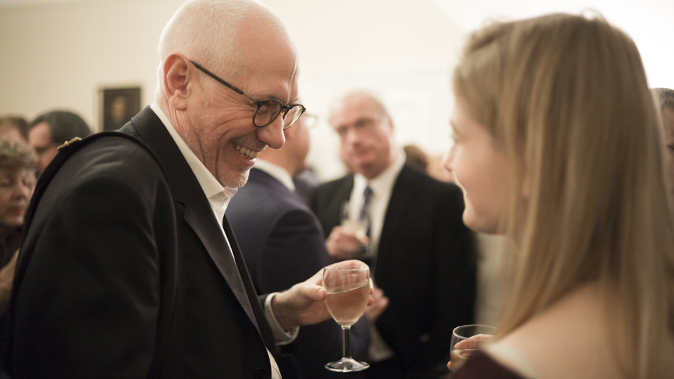 A man with glasses, holding a glass, smiling at a women with long blonde hair, also holding a glass, in a social reception.