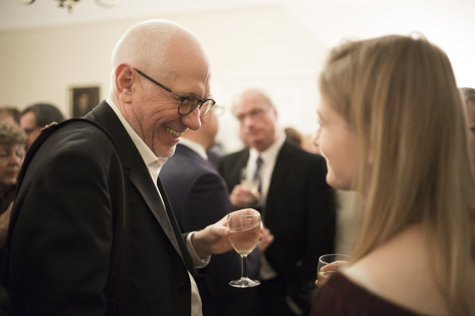 A man with glasses, holding a glass, smiling at a women with long blonde hair, also holding a glass, in a social reception.