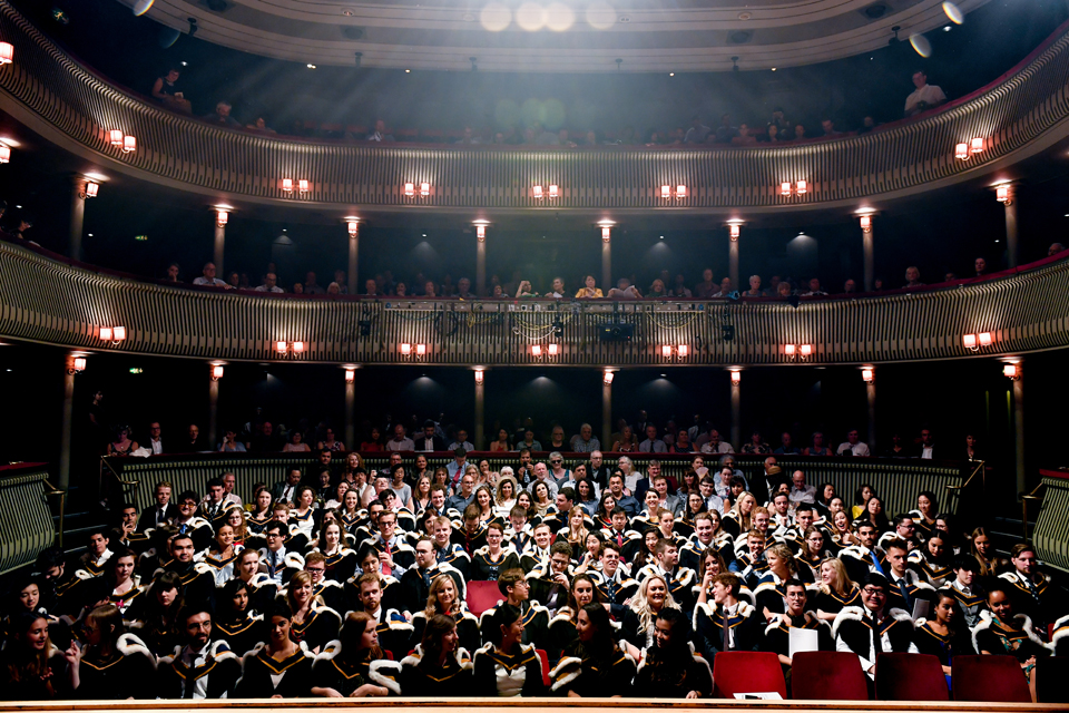 A group or graduating students, smiling at the camera, sitting in red seats, sitting in a theatre.