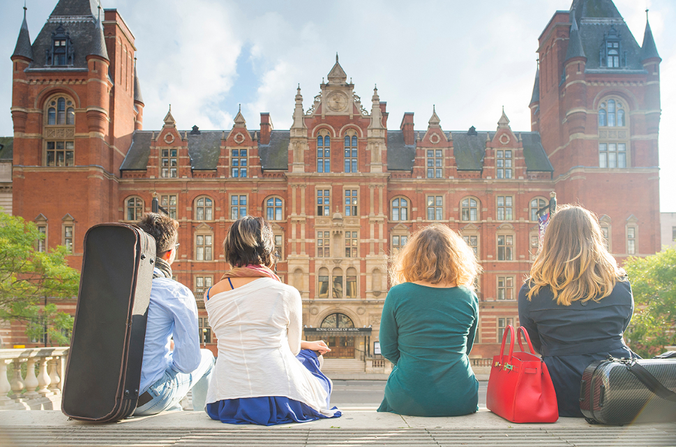 A group of students of students, with bags and music cases next to them, facing the front of the Royal College of Music.