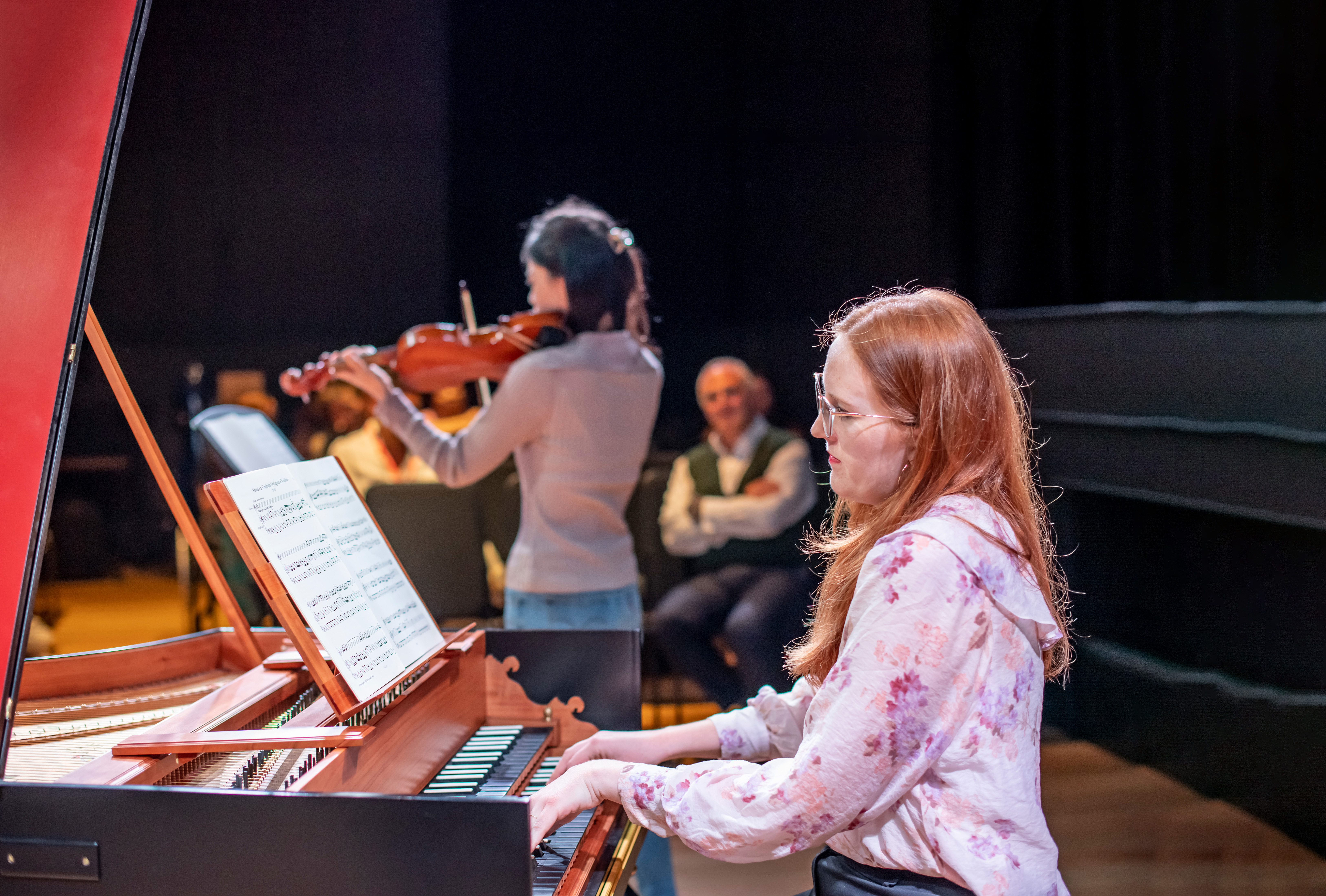 A female student, with glasses, red hair and a pink shirt, playing on a harpsichord, with a student performing in the background. A female student, with glasses, red hair and a pink shirt, playing on a harpsichord, with a student performing in the background.