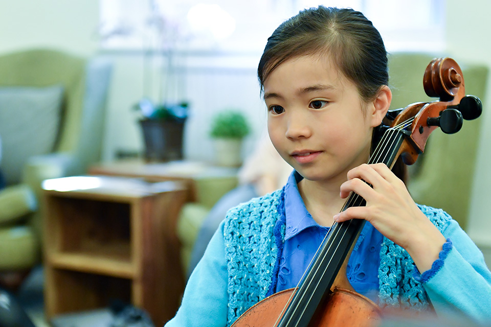 A young girl with dark hair, playing the cello, in a room green armchairs.