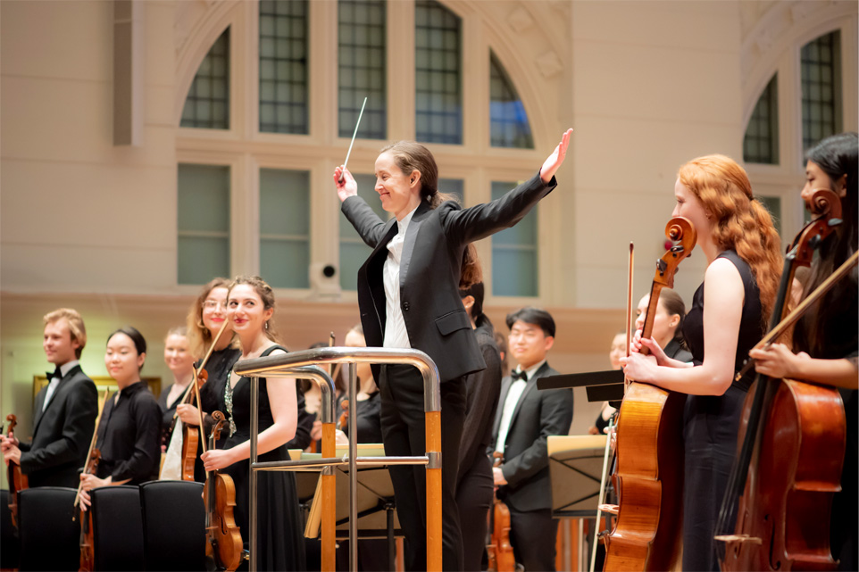 A women, with brown hair tied back, wearing formal attire and smiling, with her arms out, standing in the conductor's podium, with students, wearing formal attire, standing next to her and smiling.