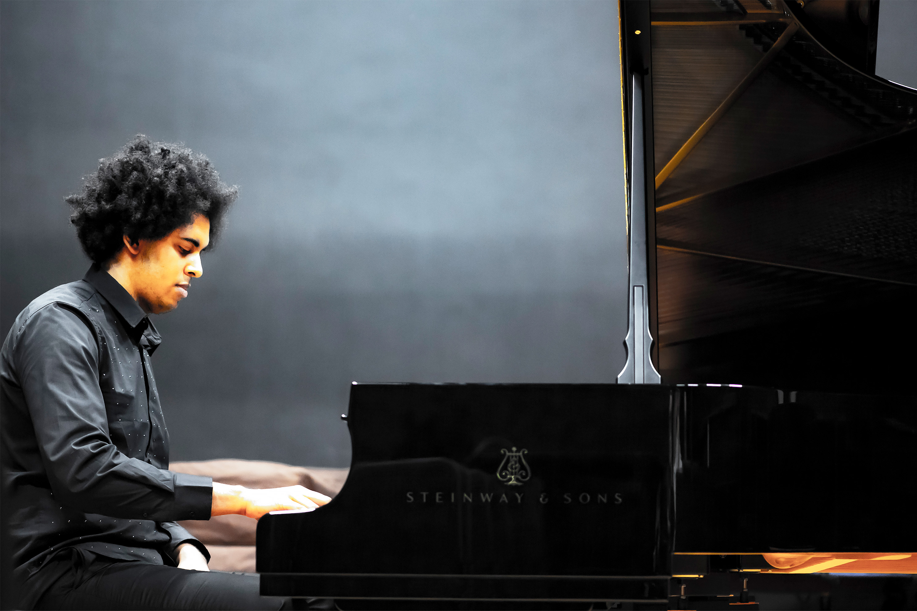 A male student playing the piano, with a dark background behind him. A male student playing the piano, with a dark background behind him.