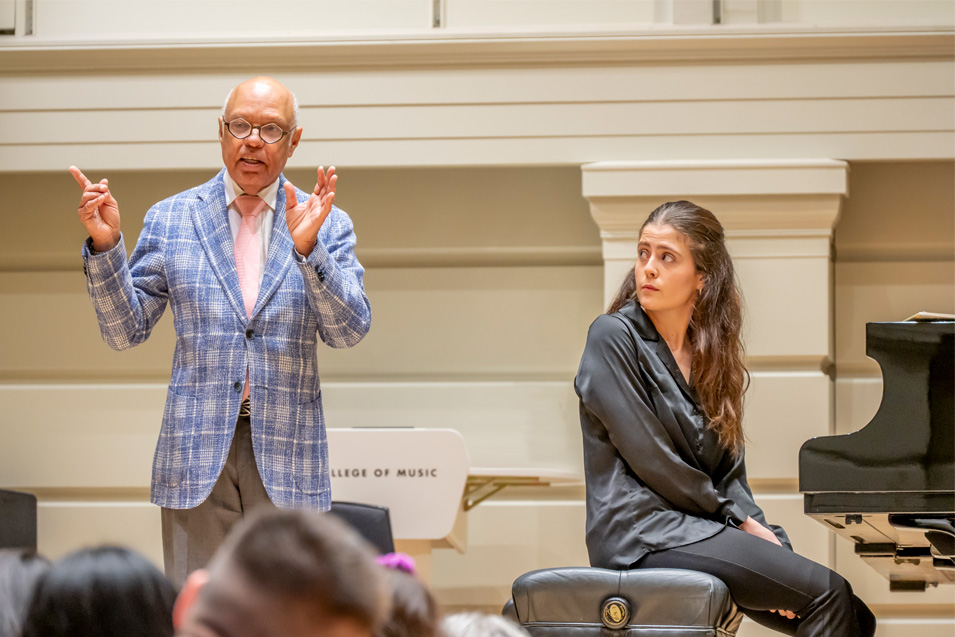 A man wearing glasses and a light blue jacket, talking to the audience, with a female student wearing smart black attire, looking and listening to him, standing on a well lit stage.