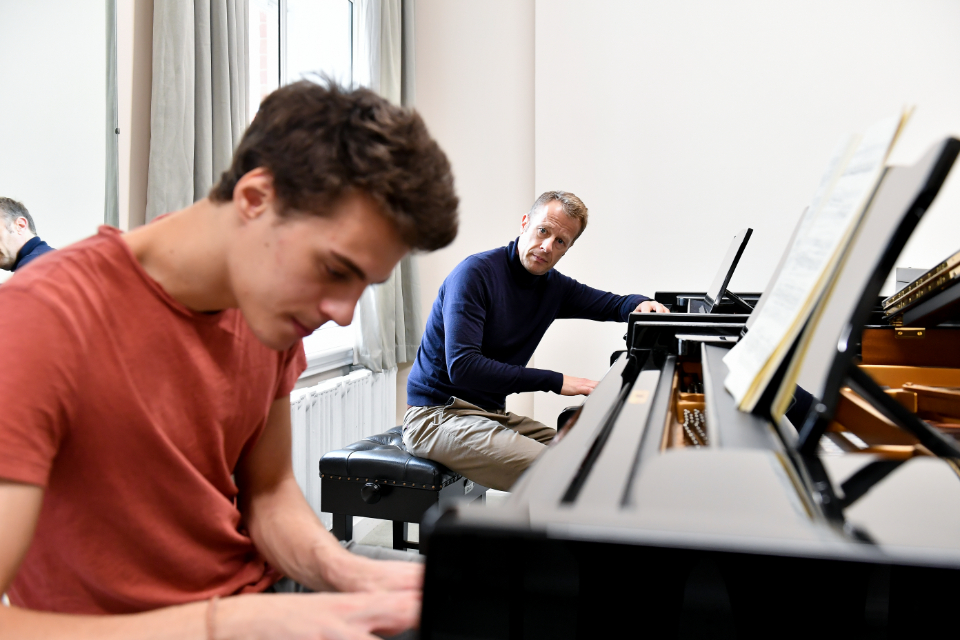 A male student performing on a keyboard, with a man wearing a dark top observing him.
