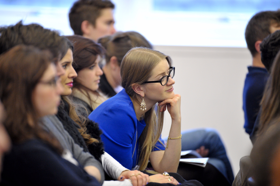 A female student with glasses, leaning in and listening, sitting with other students.