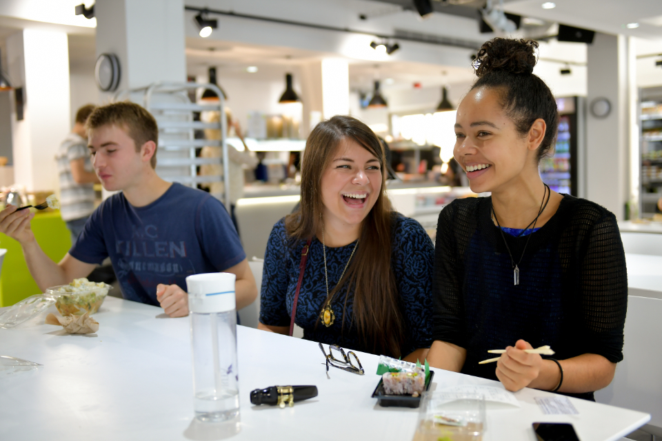 Three students smiling and chatting in a cafe, with some food, water bottles and their glasses on the table.