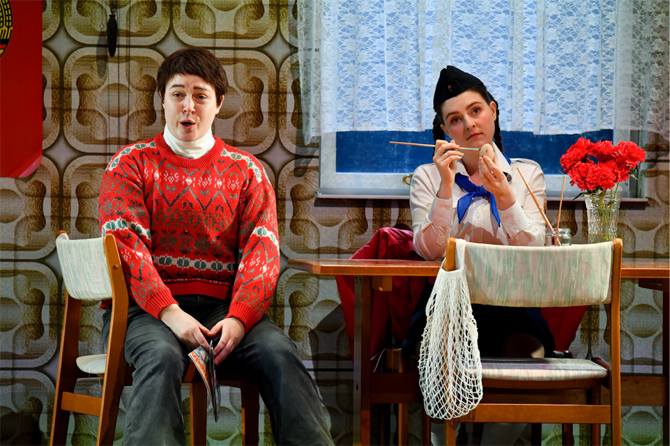 A male singer, wearing a red jumper, sitting at a wooden dining table, with a woman, dressed in a blouse with a blue ribbon, performing an opera scene on stage.