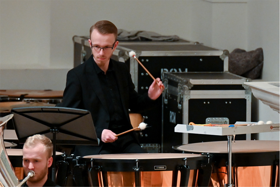 A man with glasses, wearing formal attire, performing timpani, in an orchestra performance.