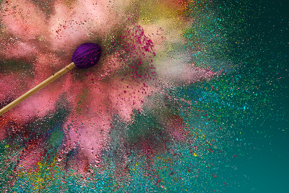 A purple drumstick surrounded by pink, gold, orange, blue and green glitter and powder, surrounded by a teal background.