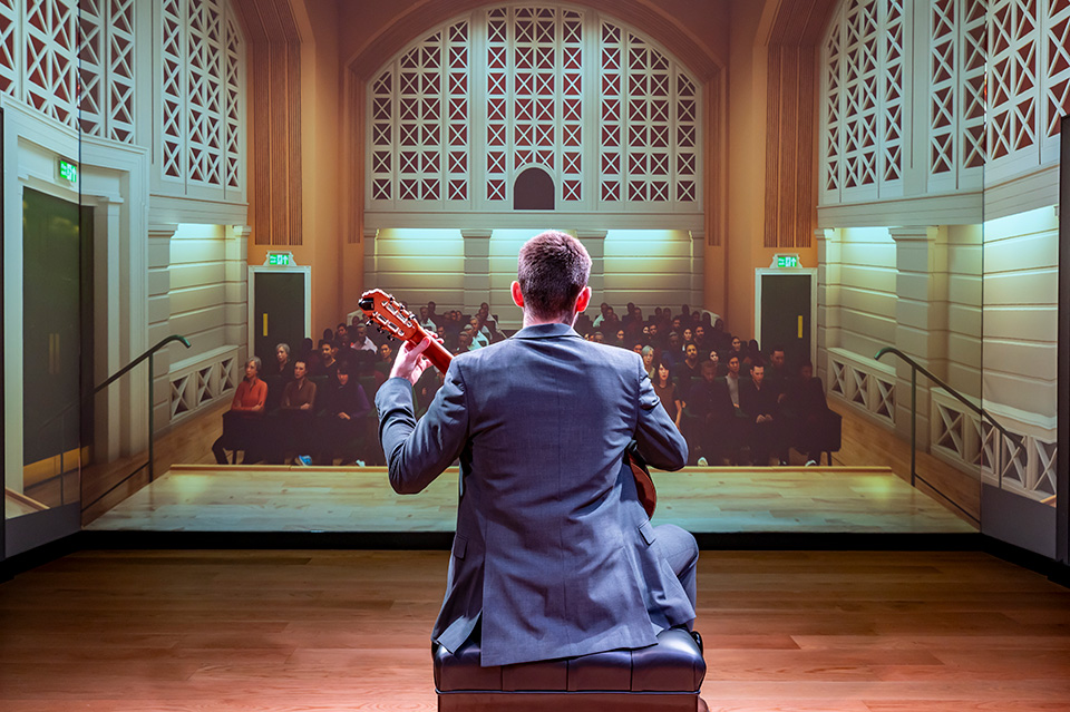 A male guitar student, wearing a smart suit, holding a guitar, performing to a virtual audience.