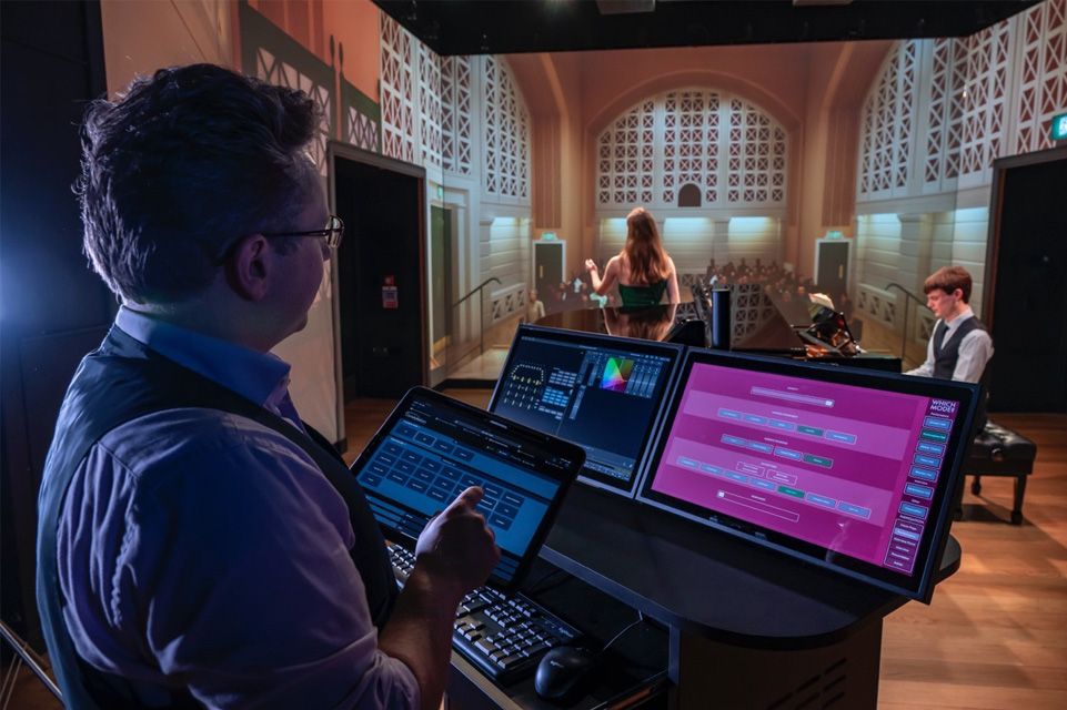 A man wearing a light blue shirt, using digital tablet to control a visual simulator, with two students, a female soprano and a male pianist, performing in front of a simulator.