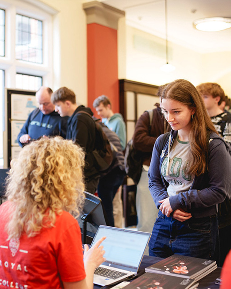 A young woman stands at a table talking to staff with RCM brochures at Open Day.