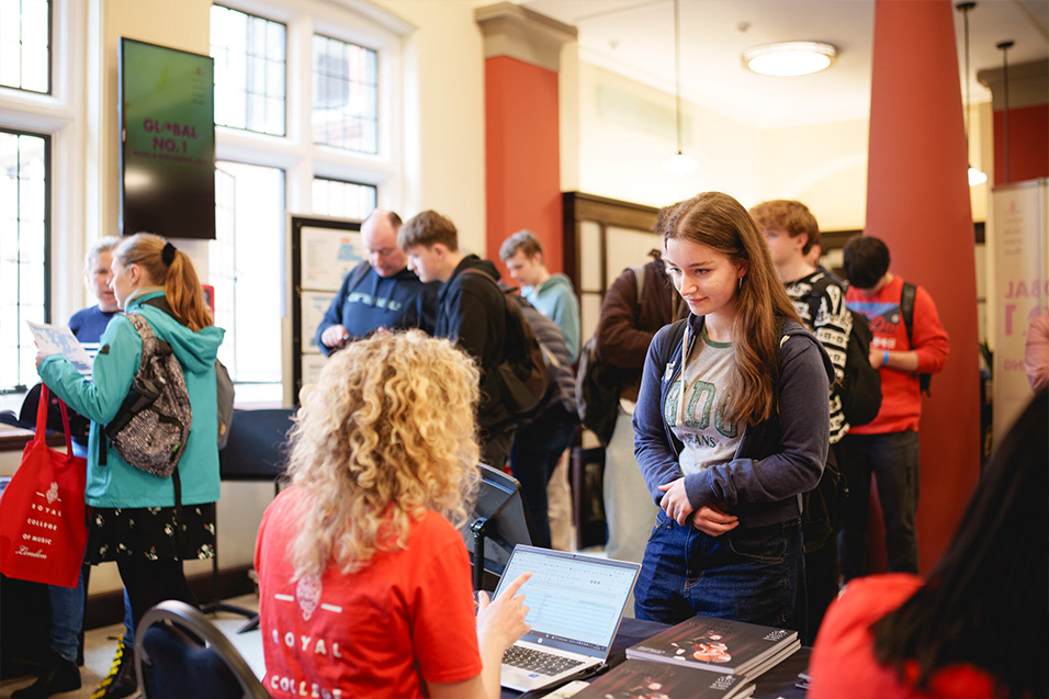 A young woman stands at a registration desk, engaging with a staff member wearing a red shirt that reads 